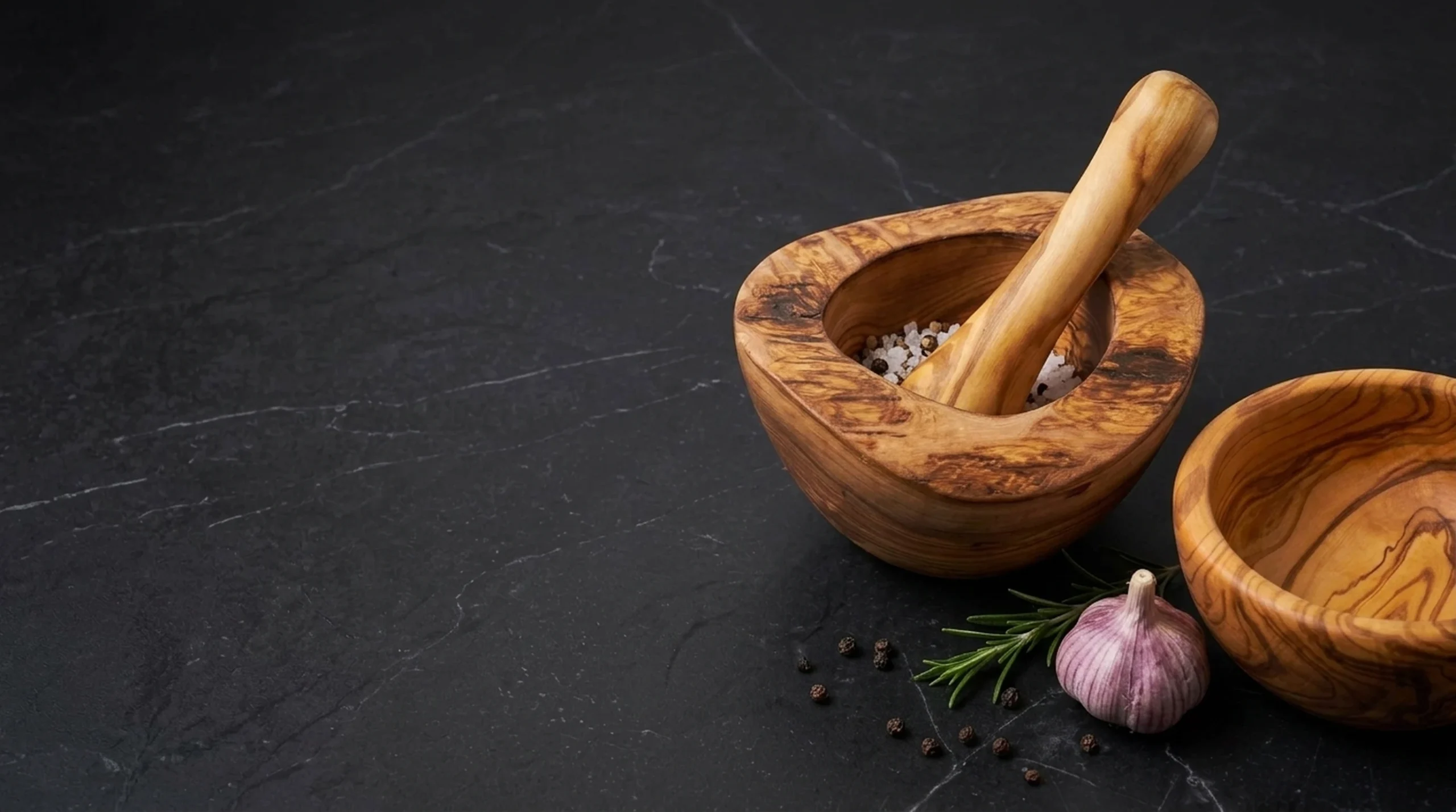 Handcrafted olive wood rustic mortar with pestle and turned bowl on dark charcoal slate with garlic and rosemary