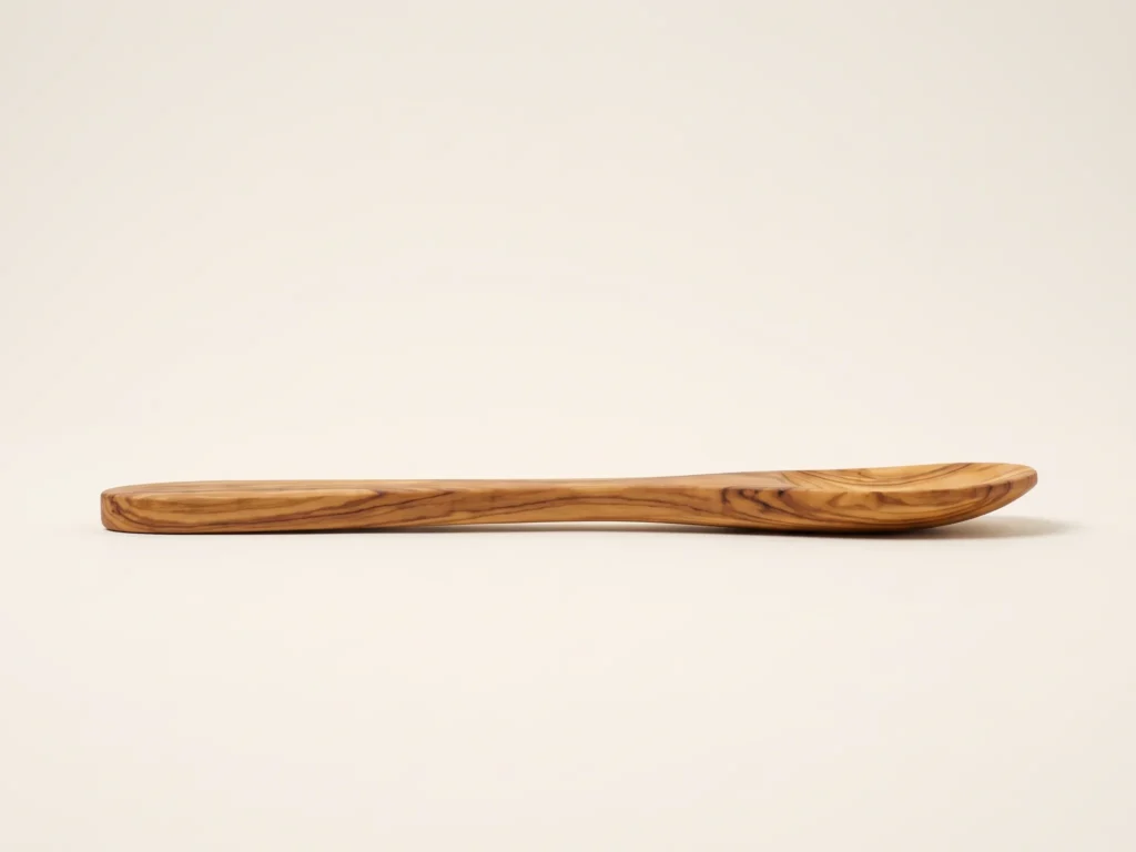 Low-angle profile grazing view of olive wood spoon documenting slim flat shape and 1.0 cm handle thickness